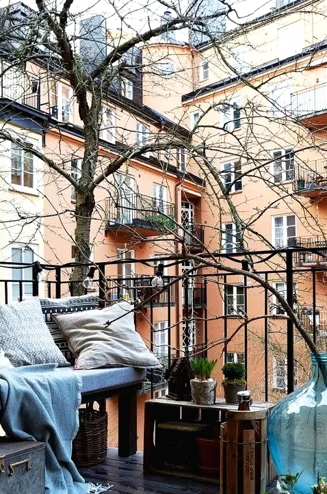 a Nordic balcony with an upholstered bench, crates and baskets, a metal chest fro storing blankets and pillows, potted greenery and lights