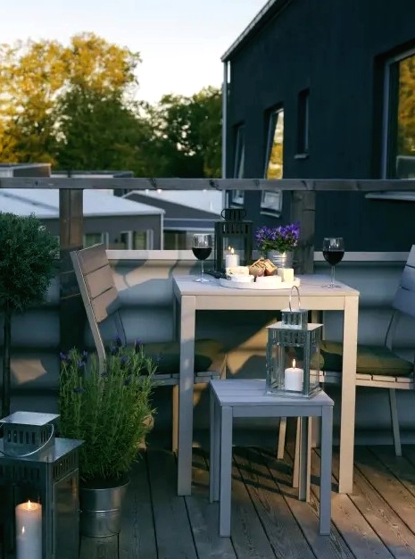 a small and cozy summer balcony with grey wooden furniture, potted greenery and blooms and candle lanterns