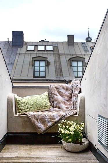 a very small Scandinavian balcony with a built-in seat with pillows and a blanket, potted blooms is a simple and chic space to stay