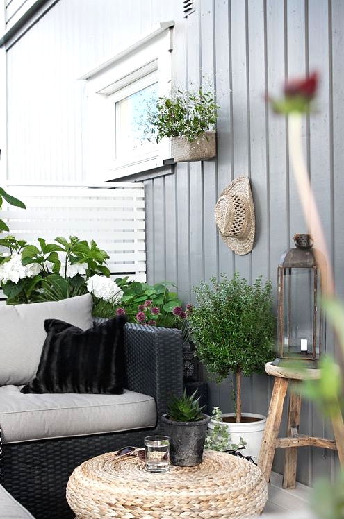 a lovely Scandinavian balcony with black wicker furniture with grey and black upholstery, a jute pouf, candle lanterns and potted blooms and plants
