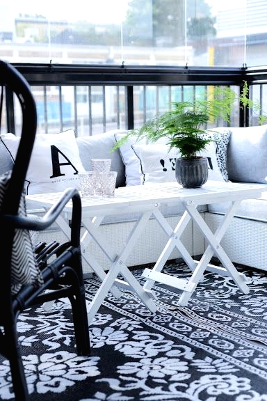 a black and white Scandinavian balcony with a printed rug, a white corner sofa, a black chair and white folding tables and greenery