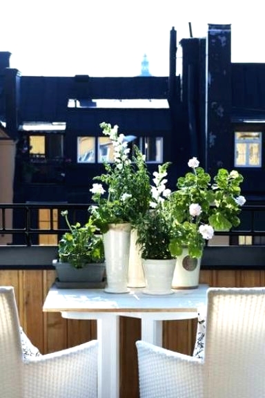 a tiny Nordic balcony with a white table and white wicker chairs, with potted blooms and greenery and a lovely view