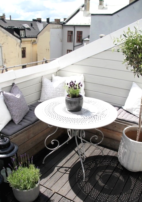 a cool Scandinavian balcony with a corner sofa with pillows and cushions, candle lanterns, potted blooms and greenery and a white table