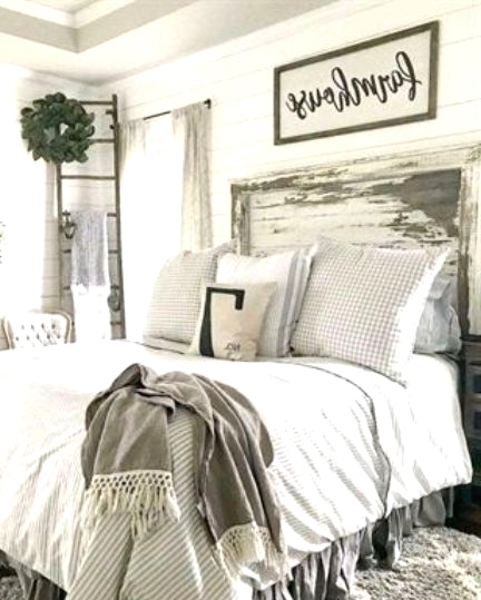 a white farmhouse bedroom with white planked walls, a whitewashed and sahbby chic headboard, neutral bedding, a ladder with a greenery wreath
