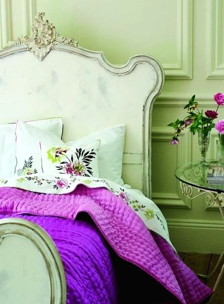 a pastel bedroom with paneling, a whitewashed vintage bed with neutral and bold purple bedding, a glass table with blooms