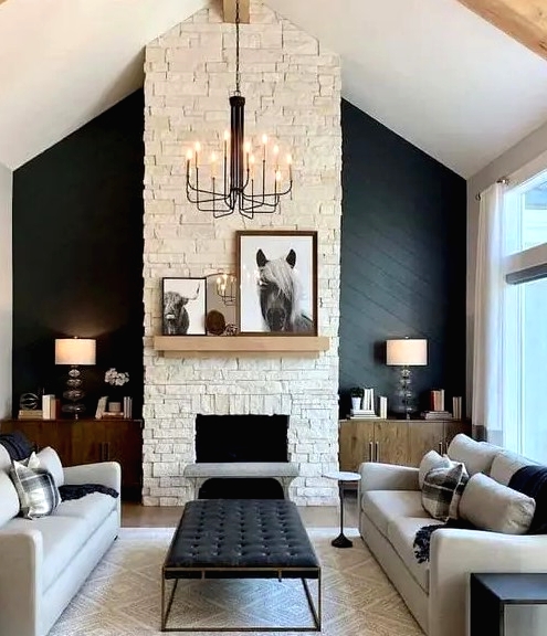 a chic living room wiер a black accent wall, a fireplace, grey sofas, black benches, a black ottoman and a vintage chandelier