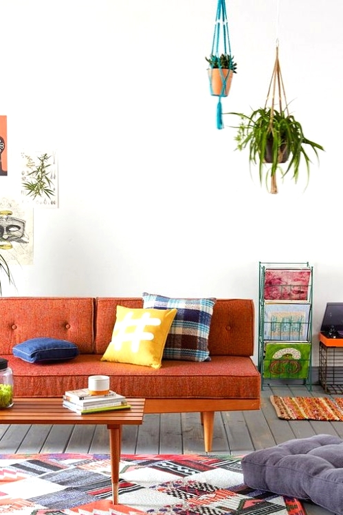 a mid-century modern to boho living room with a rust-colored sofa and bold pillows, a bright rug, potted greenery and shelving units with magazines