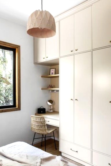 a neutral tan bedroom with a bed with neutral bedding, a large creamy wardrobe and a built-in vanity and open shelves, a plywood pendant lamp