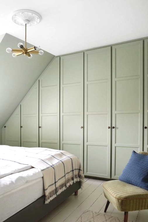 a small attic bedroom with a series of green wardrobes, a grey bed and a green chair plus a gold chandelier