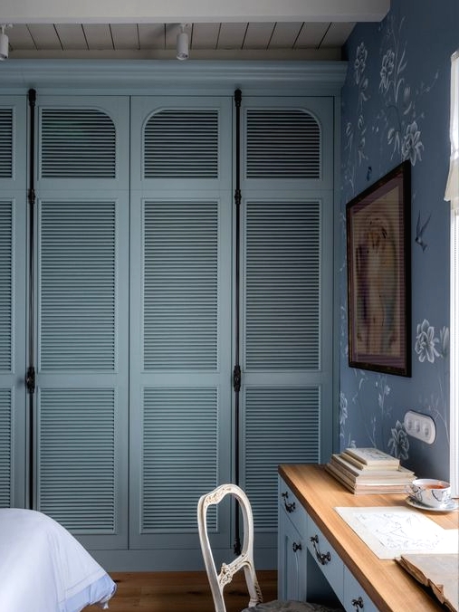 a vintage bedroom with a blue and white printed wallpaper wall, a blue desk and a vintage chair, a wall-to-wall wardrobe with blue shutter doors, a bed with bedding