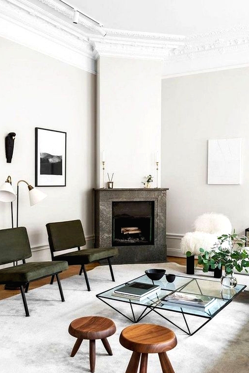 a refined living room with a stone fireplace, a glass coffee table with a geometric base, wooden stools and dark green leather chairs