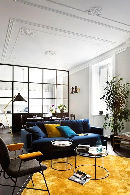 an elegant living room with a navy sofa, a black and yellow chair, a yellow hexagon-shaped rug and pillows plus potted plants