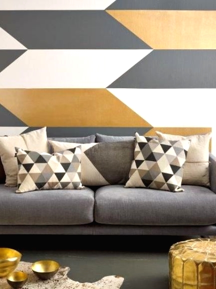 an elegant modern living room with a chevron printed accent wall, a grey sofa with geo printed pillows, a gold pouf and a neutral rug with gold bowls