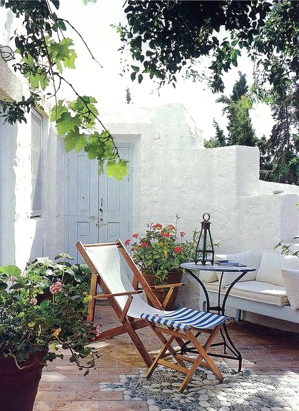 a little Mediterrranean outdoor nook with a sofa with white cushions and pillows, folding chairs, a metal table, bright blooms in planters