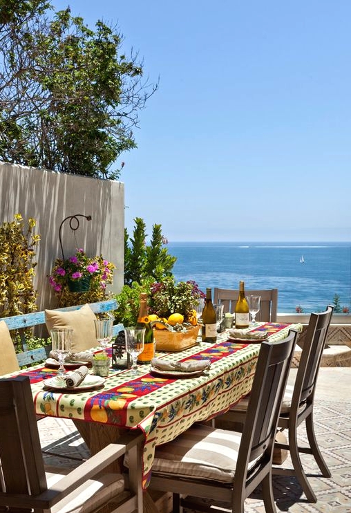 a lovely Mediterranean dining space with a view, a wooden table and chairs, a blue bench, a bright tablecloth and bright blooms