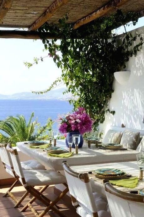 a Mediterranean dining space with a view, with a built-in sofa, white tables and folding chairs with white upholstery, blooms and greenery