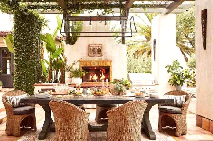 a refined Mediterranean outdoor dining space with greenery and potted plants, a dark-stained table and wicker chairs, a pendant lamp and a fireplace