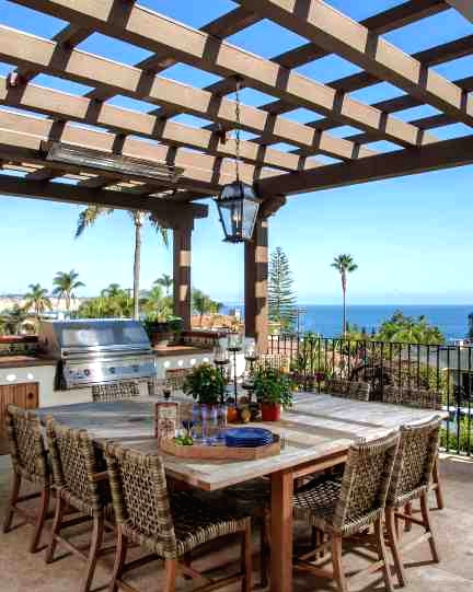 an outdoor Mediterranean dining space with a large table, wicker chairs, a pendant lamp and an outdoor kitchen plus a fantastic view