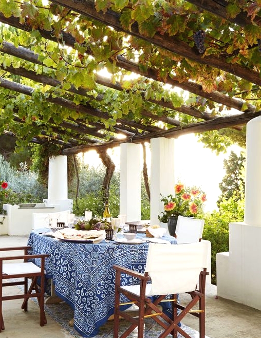 an exquisite Mediterranean patio with greenery over the space, a table with blue tablecloth and dark chairs with white upholstery and blooms