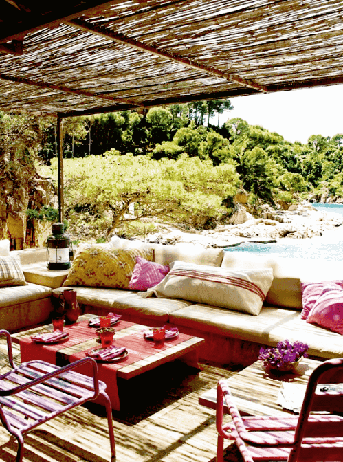 a bright Mediterranean outdoor space with a built-in upholstered bench, low tables, pink pillows, pink metal chairs and a beautiful view