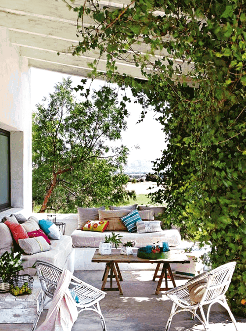 a lively Mediterranean outdoor space with a white built-in sofa with neutral upholstery and bright pillows, a wooden trestle table and wicker chairs, greenery weaving around the pillars