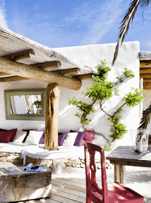 a pretty Mediterranean outdoor space with a roof over a built-in sofa with white upholstery, a wooden side table, a wooden table and a pink chair, some greenery