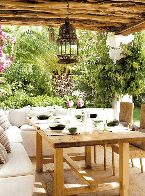 a white Mediterranean outdoor space with a built-in white sofa, a wooden table, woven chairs, a Moroccan pendant lamp and bright blooms around