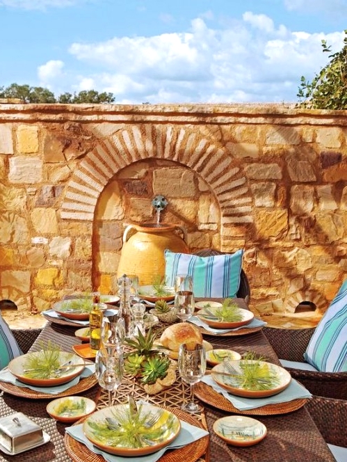 a gorgeous Mediterranean outdoor dining space with a stone wall with a fountain, a dark wicker dining seat, blue pillows, greenery and succulents for a fresh touch
