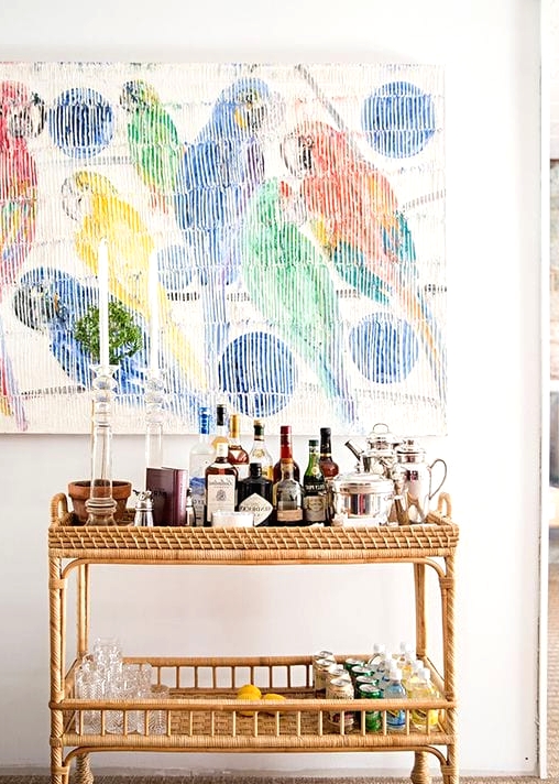 a bright space with a colorful artwork, a wicker home bar cart with lots of drinks, bottles, glasses and other stuff