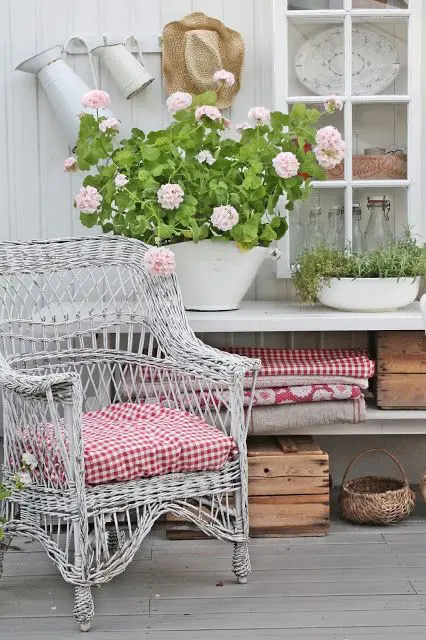 a small outdoor space with a whitewashed floor, a grey wicker chair with plaid upholstery, potted greenery and blooms, baskets and crates for storage
