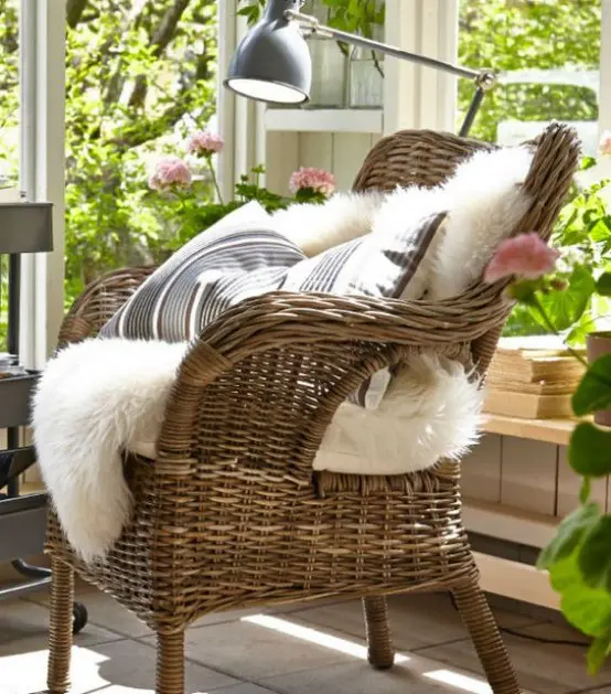 a beautiful wicker chair with neutral faux fur, printed pillows, a table lamp, blooms and greenery aorund for a welcoming reading nook