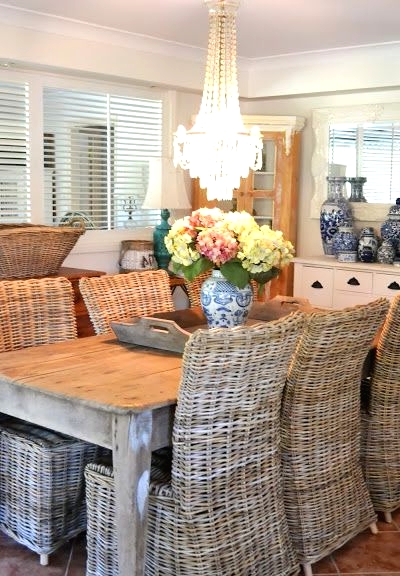 a farmhouse dining space with a wooden table, wicker chairs, blooms in a vase and a statement crystal chandelier