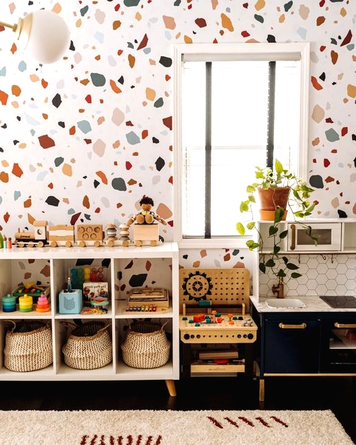 a bright mid-century modern nursery with a terrazzo accent wall, a white shelving unit, a navy play kitchen, some greenery and bright toys