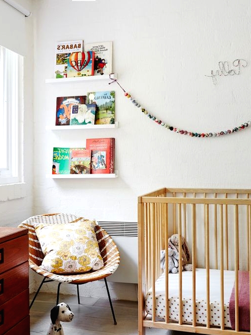 a mid-century modern nursery with open shelves for books, a stained crib, a stained dresser, a wicker round chair, printed textiles and lovely toys