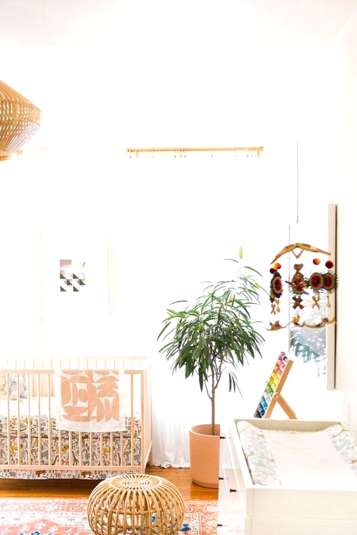 a modern boho nursery with a wodoen crib, a printed rug and bedding, a wooden pendant lamp and a stool, a white dresser, a potted plant