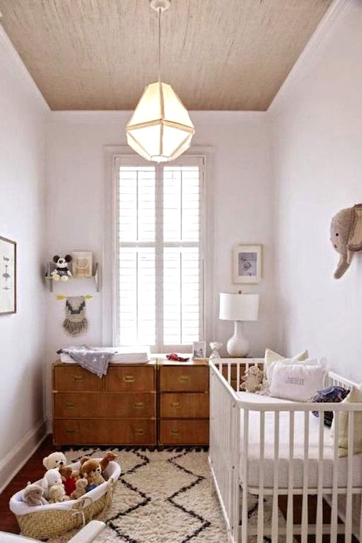 a small modern nursery in neutrals, with a white crib, a stained dresser as a changing table, a basket with toys, a table lamp and artworks