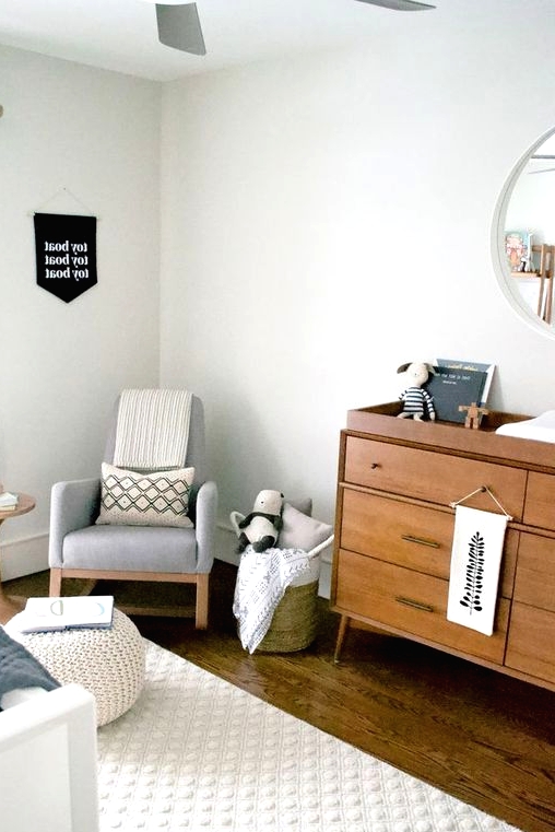 a welcoming mid-century modern nursery in neutrals, with a grey rocker, a stained dresser, a white crib, a white pouf, baskets with toys and printed textiles