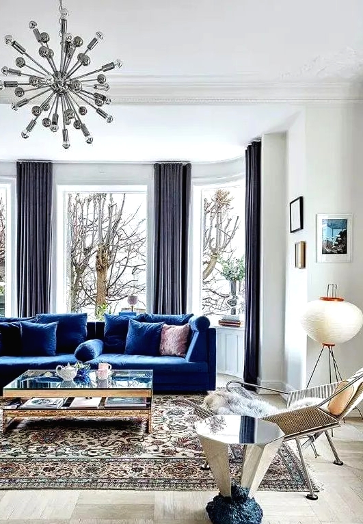 a light-filled living room wiht a bay window, a bold blue sofa, a glass coffee table and a mirror side table, a cool chair and a mid-century modern printed rug