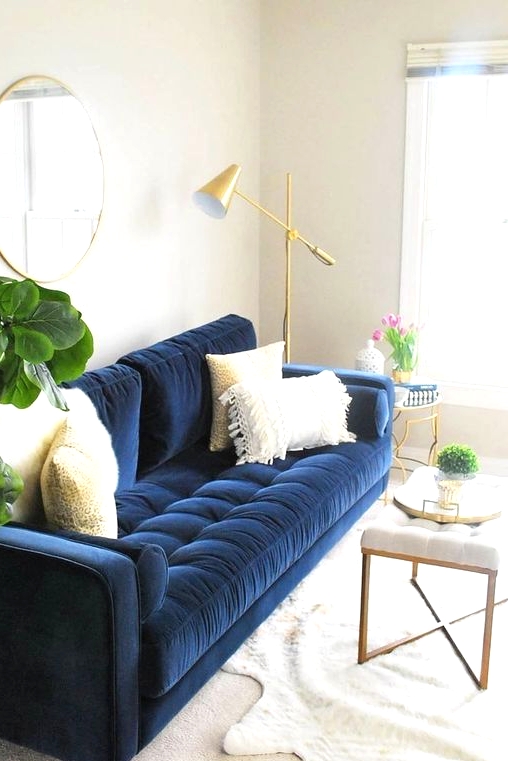 a neutral living room with a navy sofa with neutral pillows, a round mirror in a gold frame, a creamy upholstered bench, a gold floor lamp and a side table