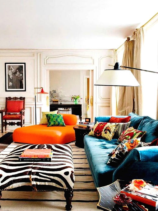 a beautiful maximalist living room with a blue sofa with colorful pillows, a zebra print ottoman, an orange daybed and cool floor lamps