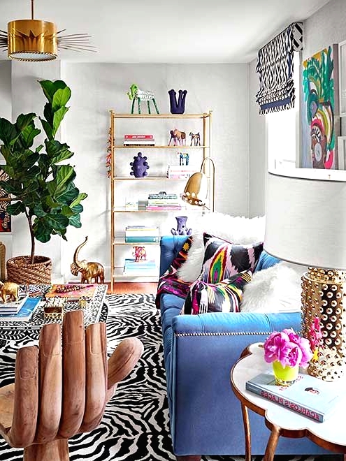 a maximalist living room with zebra printed rugs, a blue sofa with colorful pillows, a printed table, a glass shelving unit and a potted tree