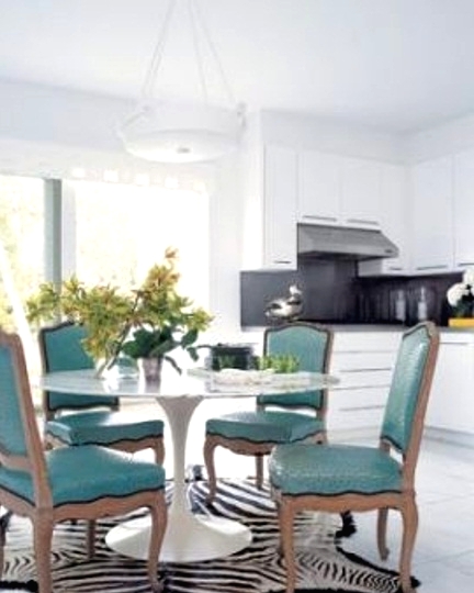 a white kitchen with a black backsplash, concrete countertops, a dining zone with a round table, a zebra print rug and turquoise chairs with a vintage design