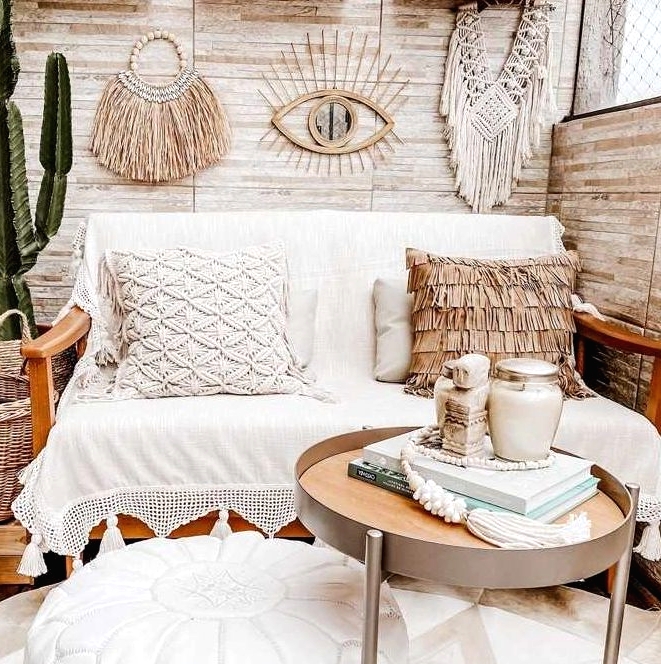 a beautiful neutral boho balcony with a sofa with boho pillows, a boho rug, a white pouf, potted plants, a side table and macrae and hangings of various kinds