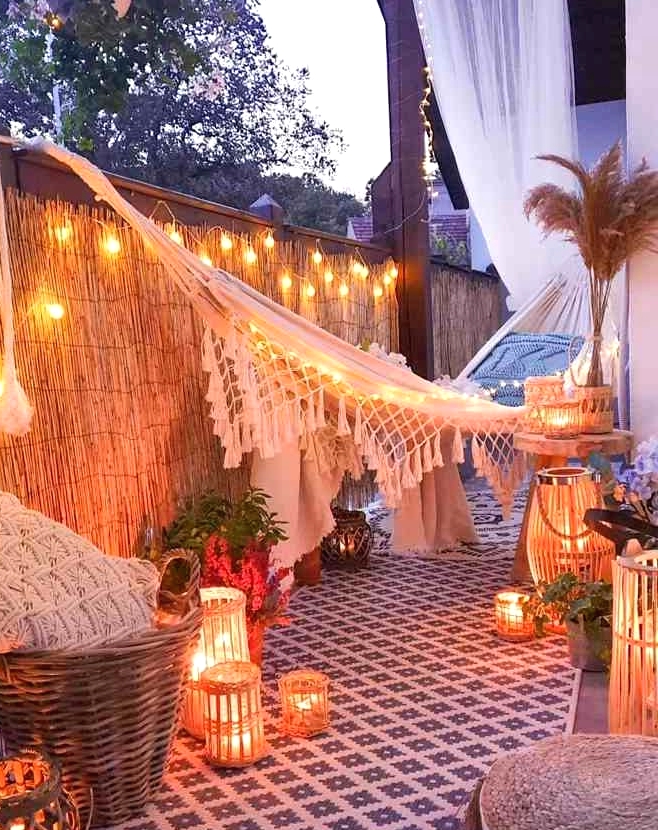 a boho balcony with an embroidered rug, lots of candle lanterns, a hammock with pillows, lamps and pillows and a jute pouf