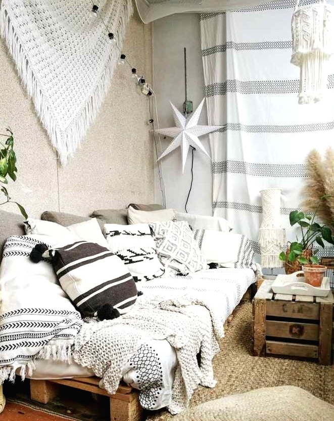 a boho balcony with a jute rug and cushions, a pallet sofa with black and white boho pillows, macrame and blankets, a crate as a table and some potted plants