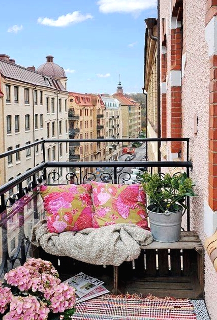 a colorful boho balcony with a bright printed pink rug and pillows on the wooden bench, some potted blooms and greenery is lovely and inviting