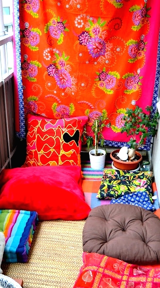 a colorful boho balcony with an orange curtain, super bright and printed pillows, a jute rug, potted greenery, a striped bold rug