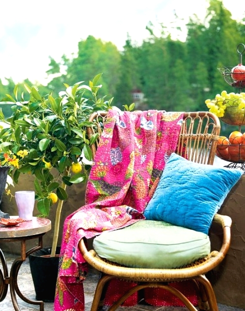 a bright and welcoming boho balcony with a rattan chair, a vintage side table, potted plants and blooms and colorful pillows and a blanket