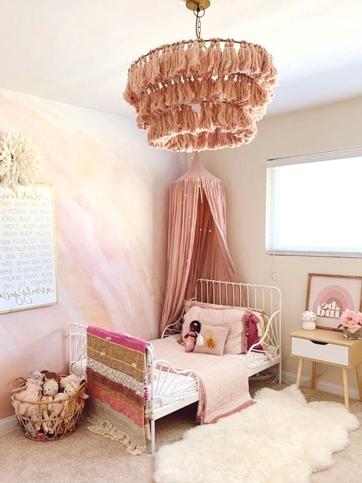 a dreamy pink girl's bedroom with a watercolor yellow and hot pink accent wall, a white metal bed with pink bedding, a pink tassel chandelier