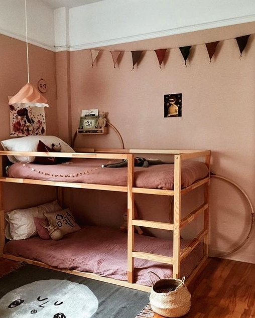 a dusty pink kid's room with dusty pink walls and bedding, an IKEA Kura bunk bed, a printed rug, a garland and baskets